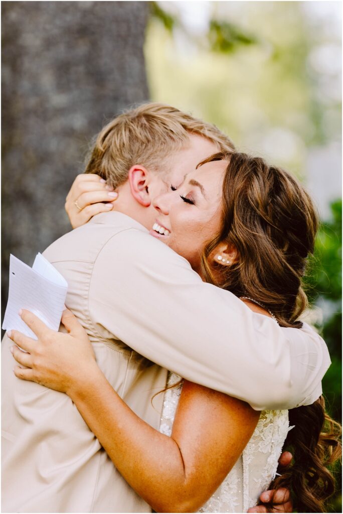 Snohomish Wedding Photographer GSquared Weddings A couple embraces outdoors, both smiling joyfully. The woman holds a piece of paper, and they are both dressed in light-colored, formal attire. Greenery and a tree trunk are visible in the background, suggesting a natural setting. Serving Seattle, Snohomish and Orlando