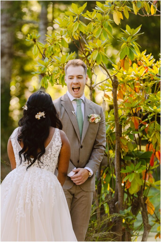 Snohomish Wedding Photographer GSquared Weddings A man in a light brown suit with a green tie looks surprised and happy as he faces a woman in a white lace dress. They are outdoors, surrounded by trees and foliage. The woman has long dark hair with a white floral hairpiece. Serving Seattle, Snohomish and Orlando