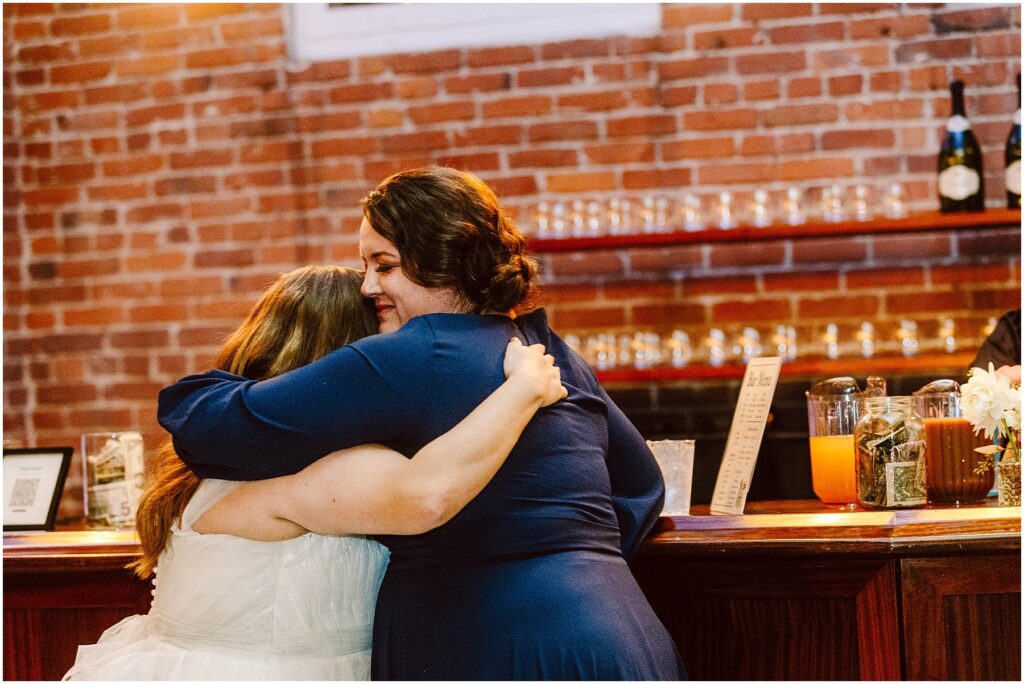 Snohomish Wedding Photographer GSquared Weddings Two women embrace warmly in front of a brick wall. One wears a white dress, the other a blue dress. Behind them is a wooden counter with glassware and drink dispensers, suggesting a celebratory event or gathering. Serving Seattle, Snohomish and Orlando