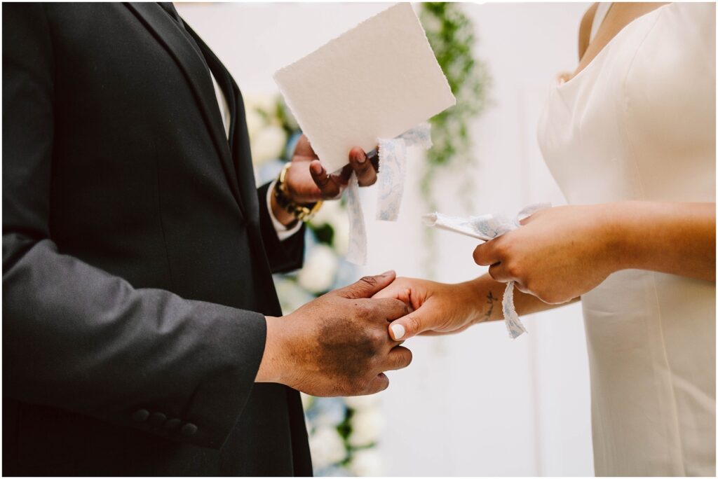 Snohomish Wedding Photographer GSquared Weddings Close-up of a wedding ceremony where a couple exchanges rings. The person on the left in a dark suit holds papers, while the person on the right in a white dress prepares to place a ring on their partners finger, with soft lighting and floral decor in the background. Serving Seattle, Snohomish and Orlando
