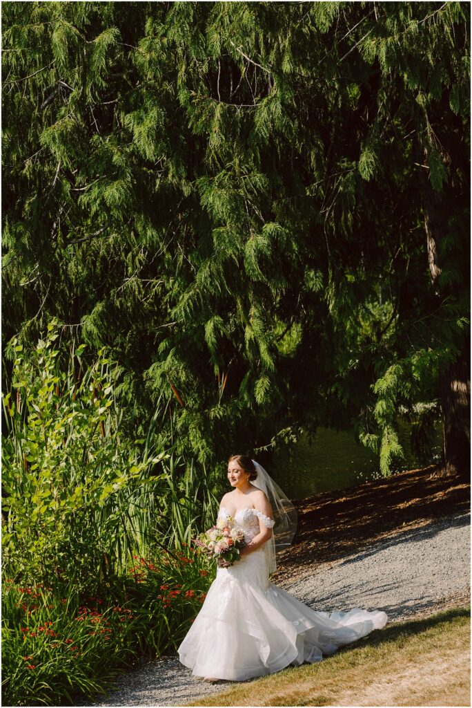 Snohomish Wedding Photographer GSquared Weddings A bride in a white wedding dress and veil walks on a gravel path, holding a bouquet of flowers. She is surrounded by lush green trees and plants, with sunlight filtering through the leaves, creating a serene outdoor setting. Serving Seattle, Snohomish and Orlando