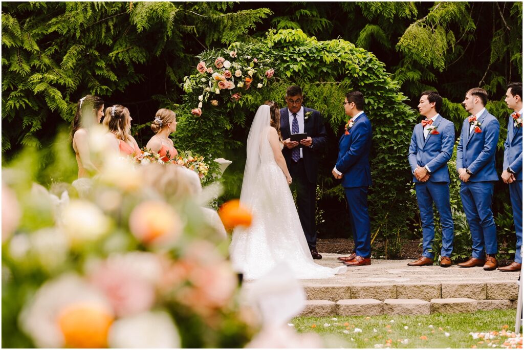 Snohomish Wedding Photographer GSquared Weddings Bride and groom stand facing each other under a floral arch during an outdoor wedding ceremony. The officiant is reading, while bridesmaids in pink and groomsmen in blue suits watch. Floral arrangements and greenery surround the scene. Serving Seattle, Snohomish and Orlando
