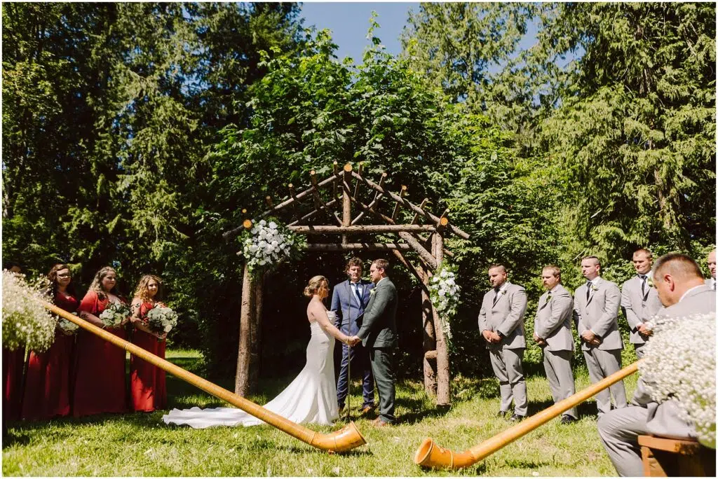 Snohomish Wedding Photographer GSquared Weddings A couple stands at a rustic wooden altar adorned with flowers in an outdoor setting surrounded by trees. Bridesmaids in red dresses and groomsmen in gray suits stand on either side. Two musicians play large wooden horns. Serving Seattle, Snohomish and Orlando