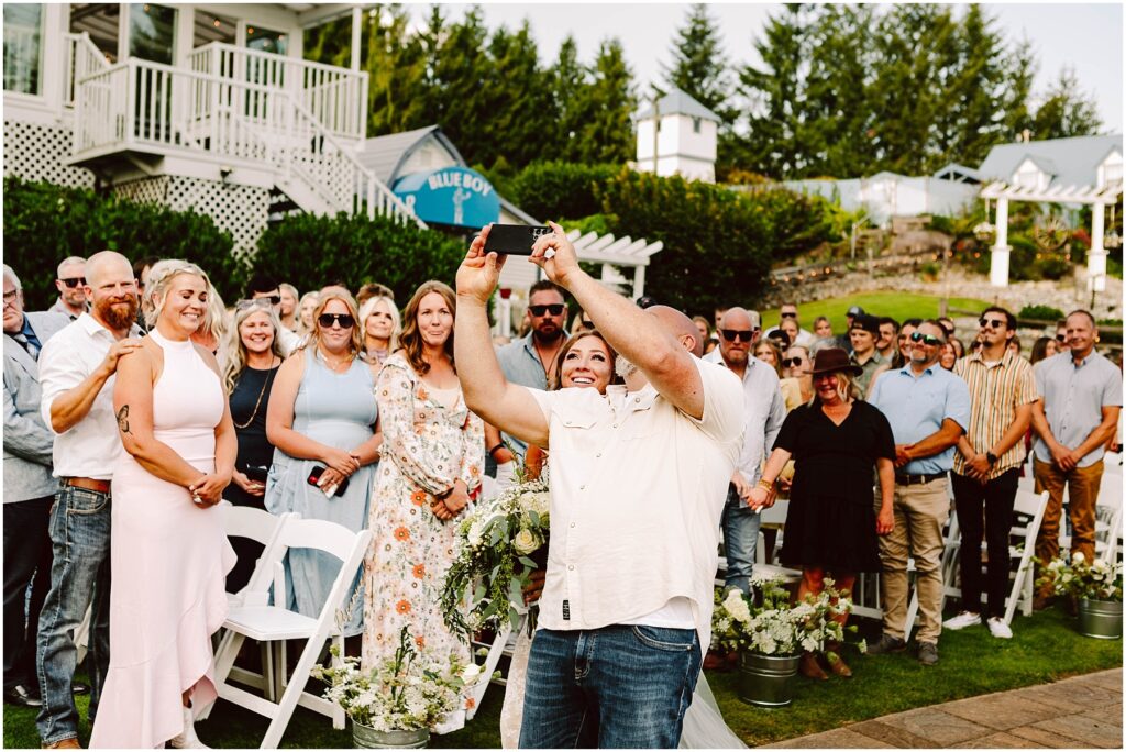 Snohomish Wedding Photographer GSquared Weddings A group of people gathered outdoors, posing for a selfie taken by a man in a white shirt. They are smiling and standing among white chairs, surrounded by greenery and a building in the background. Serving Seattle, Snohomish and Orlando