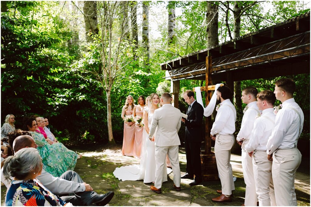 Snohomish Wedding Photographer GSquared Weddings An outdoor wedding ceremony in a lush, green setting. The bride and groom stand under a wooden pergola, with bridesmaids in peach dresses and groomsmen in white shirts and khakis nearby. Guests are seated, watching attentively. Serving Seattle, Snohomish and Orlando