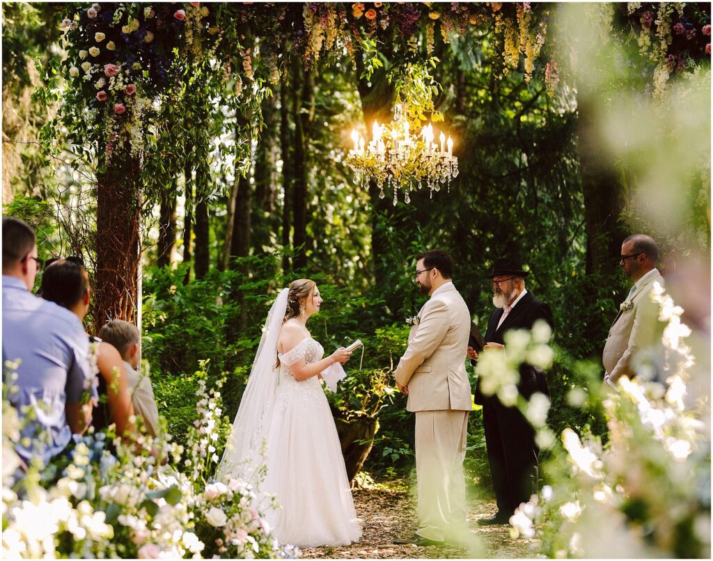 Snohomish Wedding Photographer GSquared Weddings A bride and groom stand under a floral arch, exchanging vows in a lush, green outdoor setting. A chandelier hangs above them. Guests and officiants are present, with sunlight filtering through the trees. Serving Seattle, Snohomish and Orlando