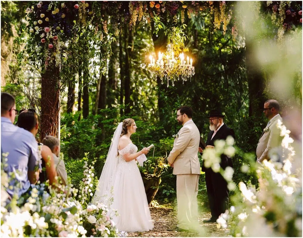 Snohomish Wedding Photographer GSquared Weddings A bride and groom stand under a floral arch, exchanging vows in a lush, green outdoor setting. A chandelier hangs above them. Guests and officiants are present, with sunlight filtering through the trees. Serving Seattle, Snohomish and Orlando