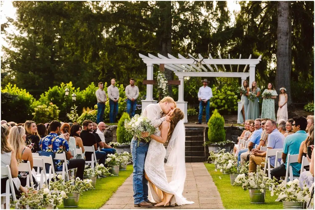 Snohomish Wedding Photographer GSquared Weddings A bride and groom share a kiss at an outdoor wedding ceremony. The groom wears a light shirt and jeans, while the bride is in a lace gown with a flowing veil. Guests are seated along both sides of the aisle, lined with flowers, under a sunny sky. Serving Seattle, Snohomish and Orlando