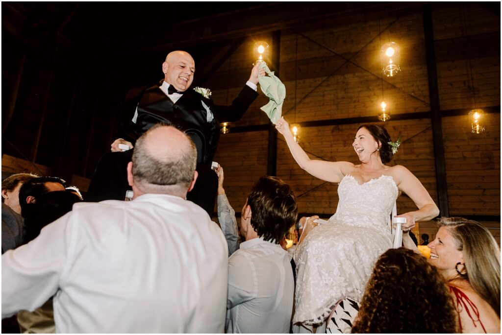 Snohomish Wedding Photographer GSquared Weddings A couple is lifted on chairs by a group of people in a warmly lit venue. The man in a suit and the woman in a white dress hold hands joyfully. Hanging lights and wooden beams are visible in the background. Serving Seattle, Snohomish and Orlando