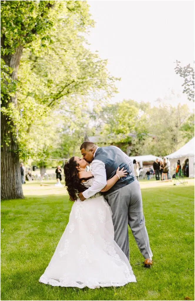 Snohomish Wedding Photographer GSquared Weddings A bride and groom share a kiss while dancing on a grassy lawn. The bride wears a white dress, and the groom is in a suit with a vest. Trees and a white tent are in the background on a sunny day. Serving Seattle, Snohomish and Orlando