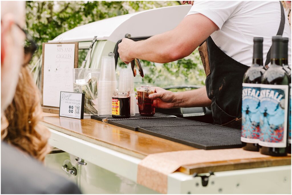 Snohomish Wedding Photographer GSquared Weddings A bartender prepares drinks at an outdoor bar set up in a vintage vehicle. The counter has cups and two bottles of wine with artistic labels. A small sign and menu are nearby. The blurred background shows greenery and a person watching. Serving Seattle, Snohomish and Orlando