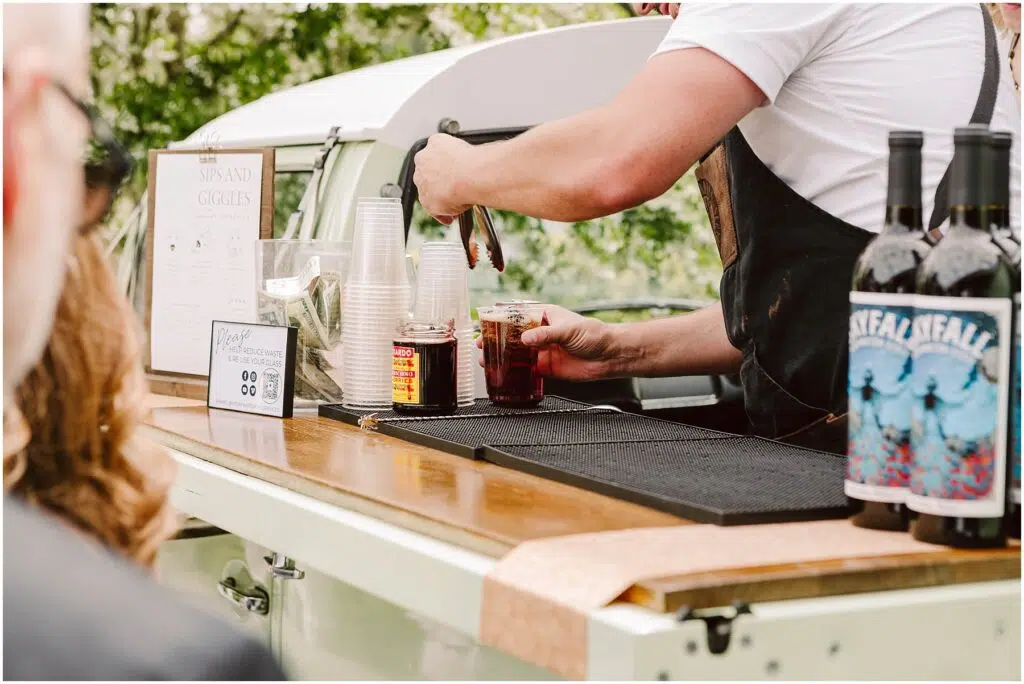 Snohomish Wedding Photographer GSquared Weddings A bartender prepares drinks at an outdoor bar set up in a vintage vehicle. The counter has cups and two bottles of wine with artistic labels. A small sign and menu are nearby. The blurred background shows greenery and a person watching. Serving Seattle, Snohomish and Orlando
