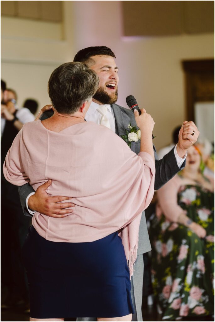 Snohomish Wedding Photographer GSquared Weddings A groom in a gray suit and a woman in a pink shawl dance closely while both hold a microphone. They appear to be singing or speaking together. The background shows seated guests, some in floral attire, at a wedding reception. Serving Seattle, Snohomish and Orlando