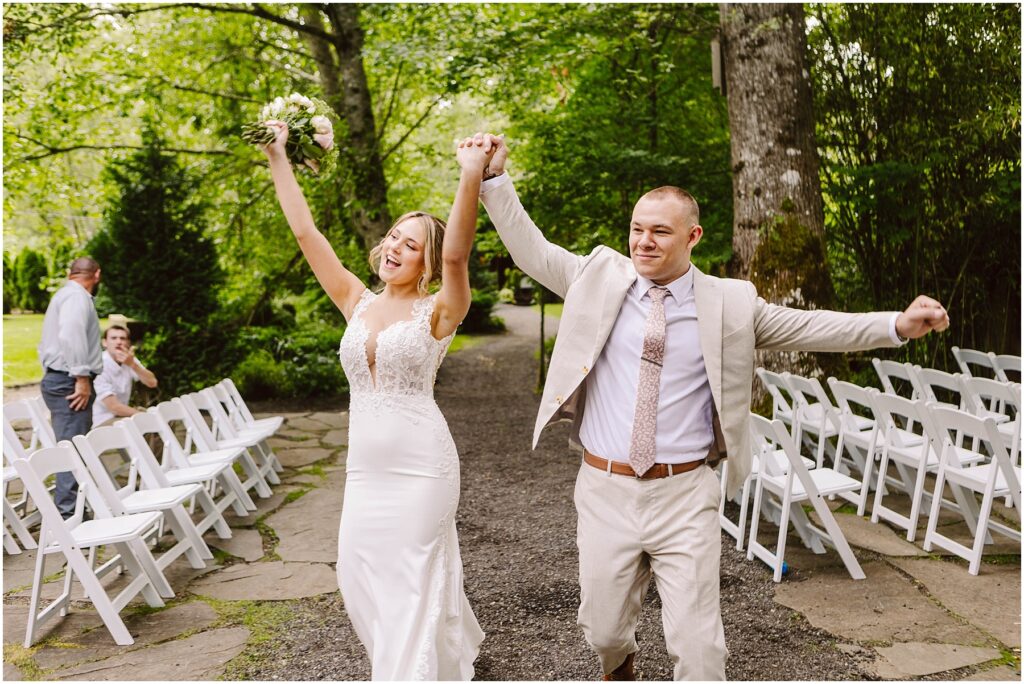 Snohomish Wedding Photographer GSquared Weddings A bride in a lace wedding dress and a groom in a light suit joyfully walk down an outdoor aisle, holding hands with arms raised. Empty white chairs line the path. Lush green trees surround the scene. Serving Seattle, Snohomish and Orlando