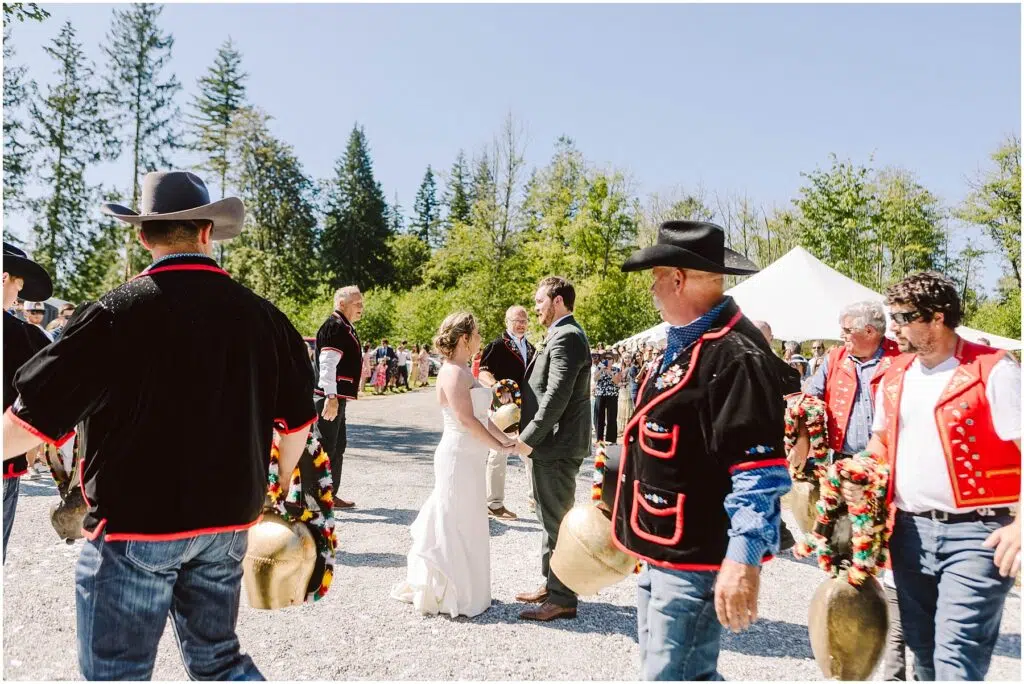 Snohomish Wedding Photographer GSquared Weddings A bride and groom stand outdoors, surrounded by men in traditional attire, some wearing cowboy hats and holding large decorated cowbells. Trees and a tent are in the background, with clear blue skies above. Serving Seattle, Snohomish and Orlando