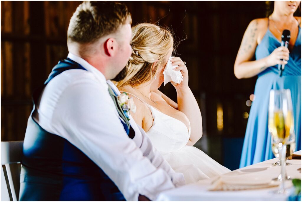 Snohomish Wedding Photographer GSquared Weddings A bride wipes her tears with a tissue while sitting next to the groom at a wedding reception. They are listening to a speech being given by a woman in a blue dress holding a microphone. Champagne glasses are on the table. Serving Seattle, Snohomish and Orlando