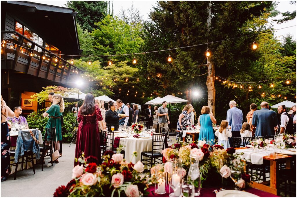 Snohomish Wedding Photographer GSquared Weddings Guests gather at an outdoor event with string lights and tables decorated with flowers. People are engaged in conversation near a building and trees, creating a warm, festive atmosphere under a clear sky. Serving Seattle, Snohomish and Orlando