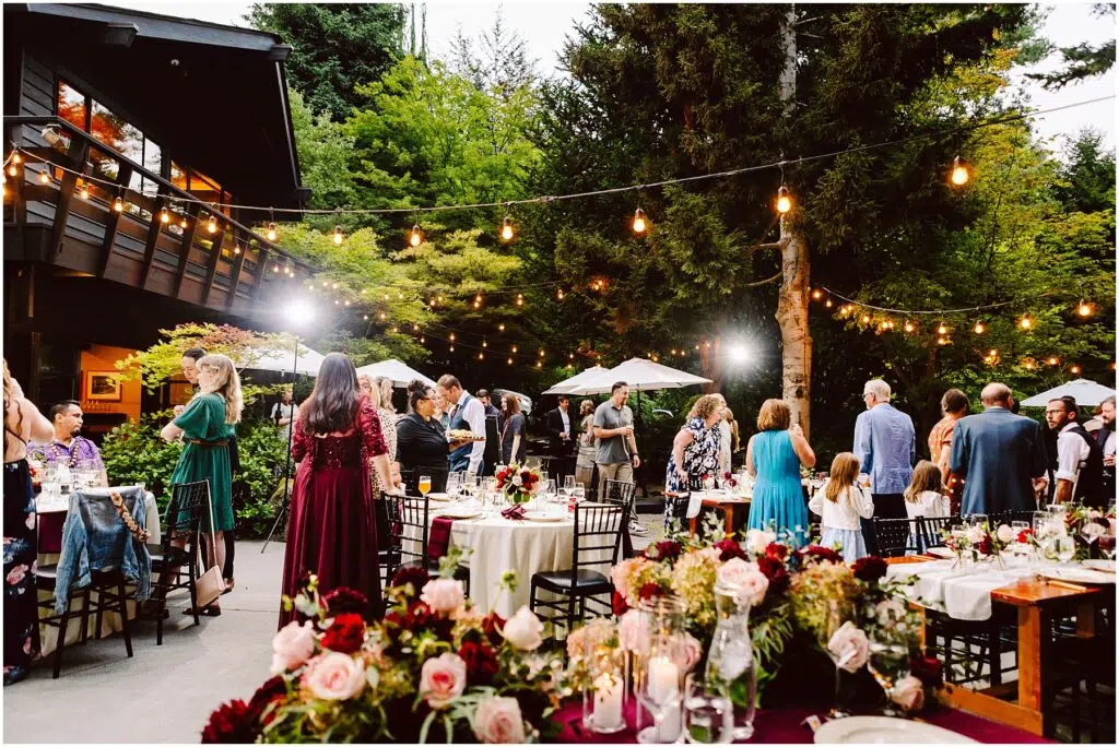 Snohomish Wedding Photographer GSquared Weddings Guests gather at an outdoor event with string lights and tables decorated with flowers. People are engaged in conversation near a building and trees, creating a warm, festive atmosphere under a clear sky. Serving Seattle, Snohomish and Orlando
