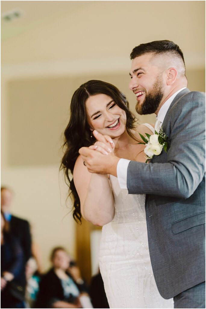 Snohomish Wedding Photographer GSquared Weddings A bride and groom joyfully dance together, smiling and holding hands. The bride wears a white gown, and the groom is in a gray suit with a boutonniere. Blurred guests are visible in the background. Serving Seattle, Snohomish and Orlando