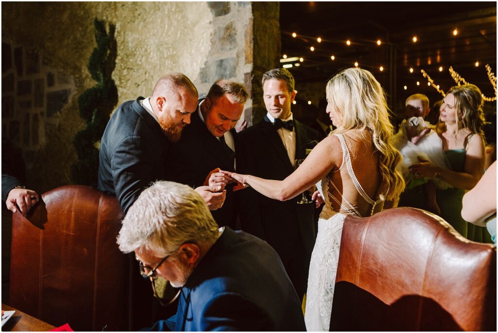 Snohomish Wedding Photographer GSquared Weddings A bride in a white gown shows her ring to three men in suits at a dimly lit event. One man leans in for a closer look, while another holds a drink. A seated older man in the foreground writes on paper. String lights hang in the background. Serving Seattle, Snohomish and Orlando