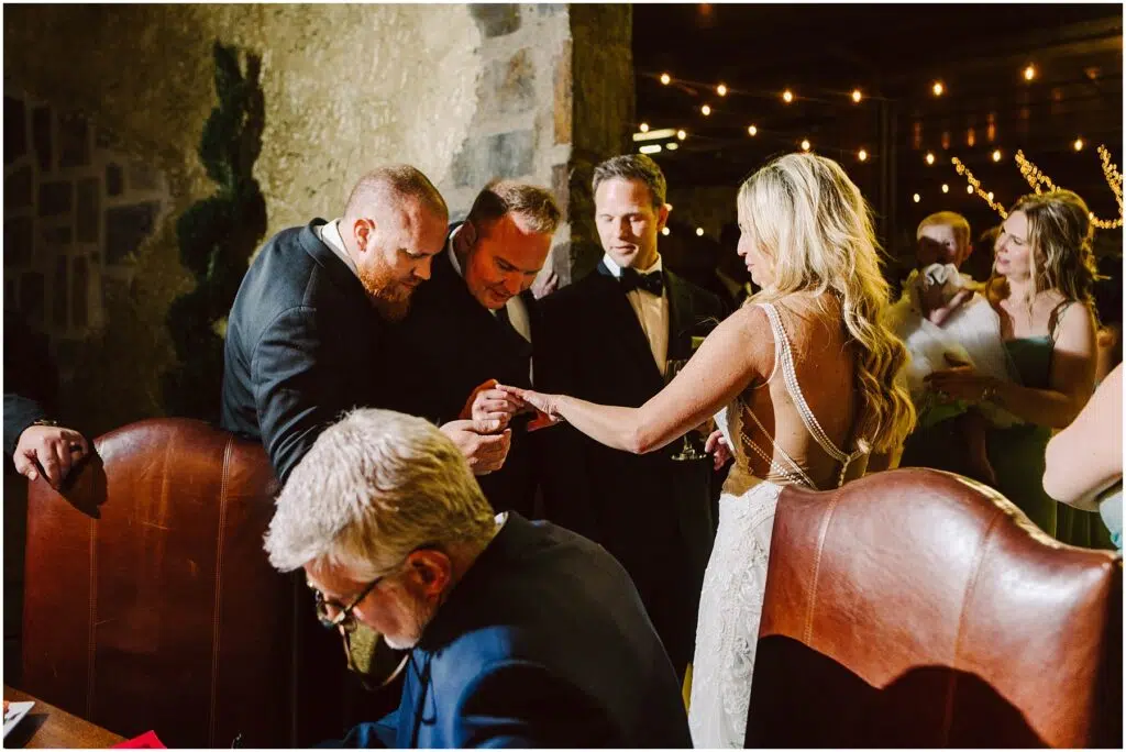Snohomish Wedding Photographer GSquared Weddings A bride in a white gown shows her ring to three men in suits at a dimly lit event. One man leans in for a closer look, while another holds a drink. A seated older man in the foreground writes on paper. String lights hang in the background. Serving Seattle, Snohomish and Orlando
