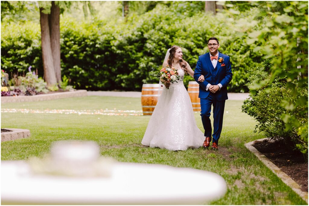 Snohomish Wedding Photographer GSquared Weddings A bride in a white gown and a groom in a blue suit walk joyfully through a lush garden. She holds a colorful bouquet, and there are wooden barrels in the background. The scene is bright and surrounded by greenery. Serving Seattle, Snohomish and Orlando