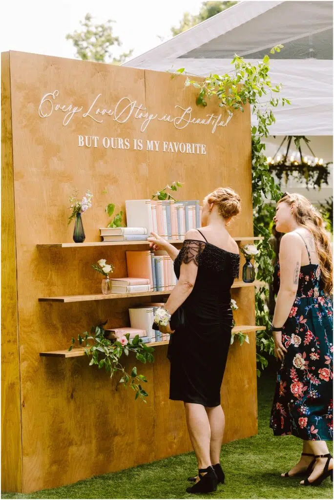 Snohomish Wedding Photographer GSquared Weddings Two women stand in front of a wooden bookshelf at an outdoor event. The bookshelf is adorned with books and plants, and a sign above reads, Every love story is beautiful, but ours is my favorite. The women are dressed in formal attire. Serving Seattle, Snohomish and Orlando