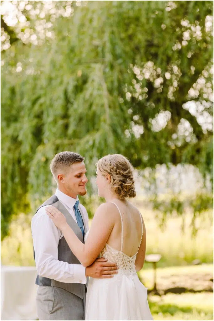 Snohomish Wedding Photographer GSquared Weddings A couple dances outdoors beneath a large tree. The woman wears a white dress with lace details, and the man wears a gray vest with a blue tie. Sunlight filters through the leaves, creating a warm, romantic atmosphere. Serving Seattle, Snohomish and Orlando