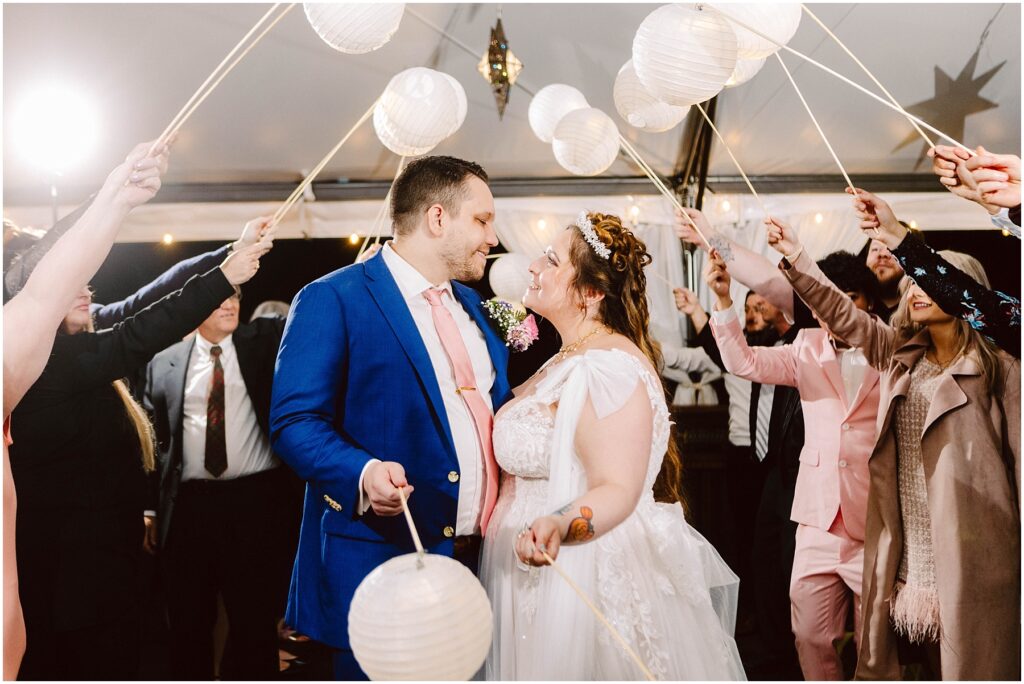 Snohomish Wedding Photographer GSquared Weddings A bride and groom smiling at each other under a canopy of white paper lanterns held by guests. The groom wears a blue suit and pink tie, while the bride is in a white dress. Some guests are dressed in suits and dresses. The setting is festive and joyful. Serving Seattle, Snohomish and Orlando