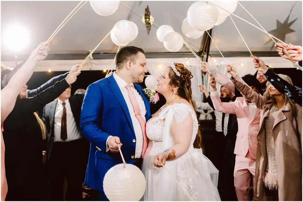 Snohomish Wedding Photographer GSquared Weddings A bride and groom smiling at each other under a canopy of white paper lanterns held by guests. The groom wears a blue suit and pink tie, while the bride is in a white dress. Some guests are dressed in suits and dresses. The setting is festive and joyful. Serving Seattle, Snohomish and Orlando