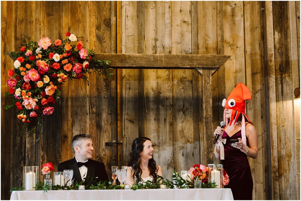 Snohomish Wedding Photographer GSquared Weddings A person wearing a squid hat stands and speaks into a microphone at a wedding reception. The couple sits at a decorated table, smiling. The backdrop is rustic wooden with a floral arrangement. Serving Seattle, Snohomish and Orlando