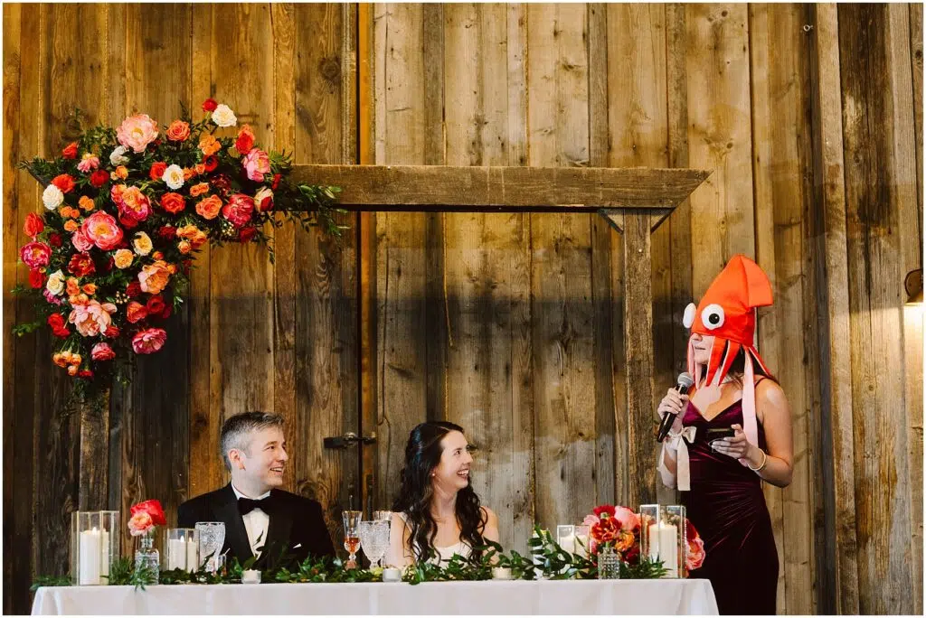 Snohomish Wedding Photographer GSquared Weddings A person wearing a squid hat stands and speaks into a microphone at a wedding reception. The couple sits at a decorated table, smiling. The backdrop is rustic wooden with a floral arrangement. Serving Seattle, Snohomish and Orlando