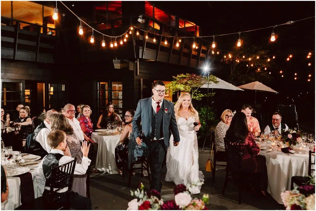 Snohomish Wedding Photographer GSquared Weddings A bride and groom hold hands, smiling as they walk through an outdoor wedding reception. Guests are seated at round tables under string lights. The evening setting includes a large building with warm lighting in the background. Serving Seattle, Snohomish and Orlando