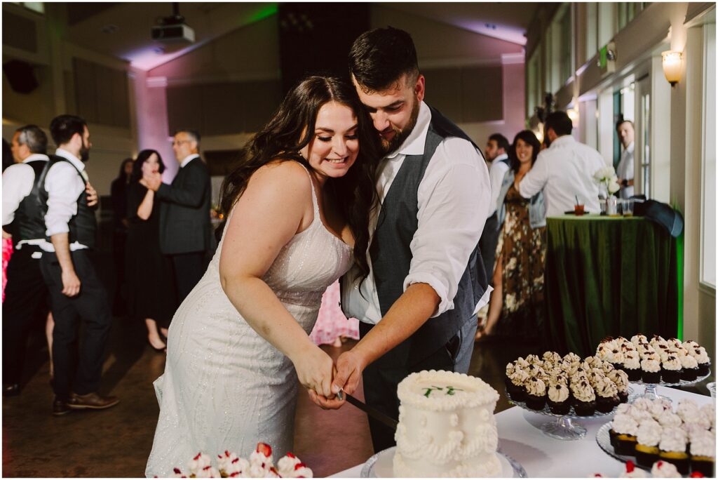 Snohomish Wedding Photographer GSquared Weddings A couple, wearing wedding attire, is smiling and cutting a small cake together at a reception. Around them, other guests are mingling, and tables are decorated with cupcakes and flowers. The room is softly lit, creating a joyful atmosphere. Serving Seattle, Snohomish and Orlando