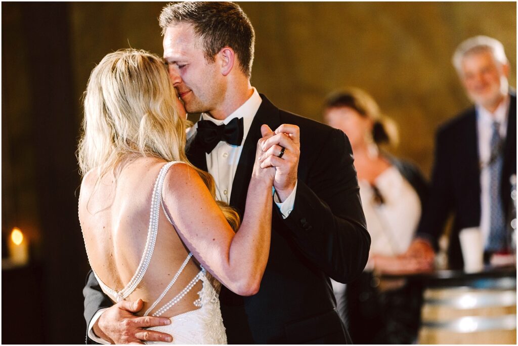 Snohomish Wedding Photographer GSquared Weddings A couple is dancing closely at an event. The woman is wearing a white dress with a low back, and the man is in a black tuxedo with a bow tie. They are surrounded by blurred onlookers, creating an intimate moment. Serving Seattle, Snohomish and Orlando