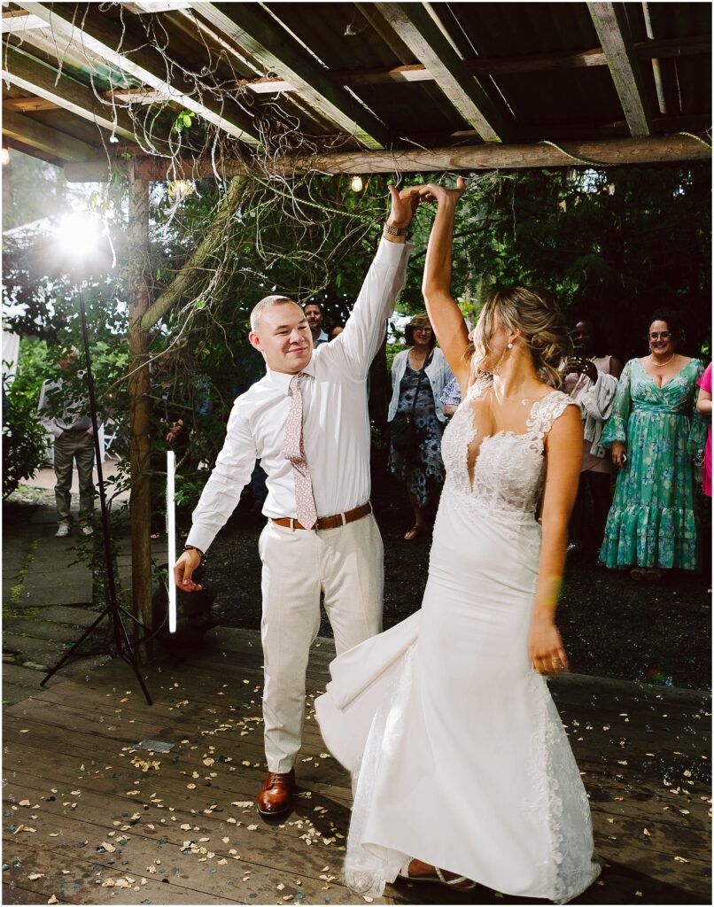 Snohomish Wedding Photographer GSquared Weddings A couple dances under a wooden pergola. The groom, in a white shirt and tie, twirls the bride, who wears a white lace wedding dress. Guests watch in the background. The setting appears to be outdoors, with greenery around. Serving Seattle, Snohomish and Orlando