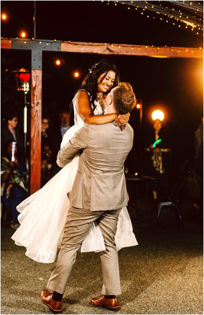 Snohomish Wedding Photographer GSquared Weddings A groom in a light gray suit lifts the smiling bride in a white dress during their outdoor wedding dance. They are surrounded by guests under warm, glowing string lights at night. Serving Seattle, Snohomish and Orlando