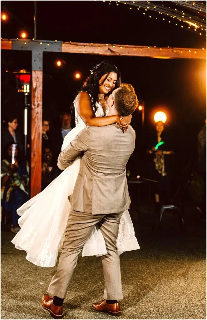 Snohomish Wedding Photographer GSquared Weddings A groom in a light gray suit lifts the smiling bride in a white dress during their outdoor wedding dance. They are surrounded by guests under warm, glowing string lights at night. Serving Seattle, Snohomish and Orlando
