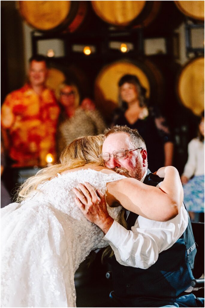 Snohomish Wedding Photographer GSquared Weddings A bride in a white lace dress is hugging an older man wearing glasses and a vest. They are in a warmly lit room with large barrels in the background. Several people are softly focused in the background. Serving Seattle, Snohomish and Orlando