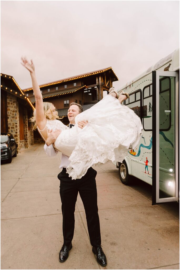 Snohomish Wedding Photographer GSquared Weddings A man in a black suit lifts a woman in a white wedding dress joyfully in front of a decorated bus. They are both smiling, and the woman has one arm raised. The setting appears to be outside, near buildings with string lights. Serving Seattle, Snohomish and Orlando