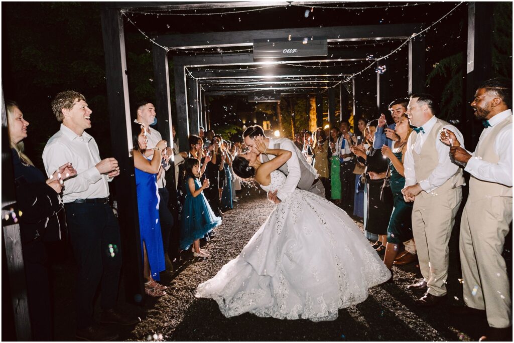 Snohomish Wedding Photographer GSquared Weddings A bride and groom share a kiss under a decorated archway at night, surrounded by applauding guests. The bride wears a white gown, and the groom is in a suit. Bubbles fill the air, adding a magical atmosphere to the scene. Serving Seattle, Snohomish and Orlando