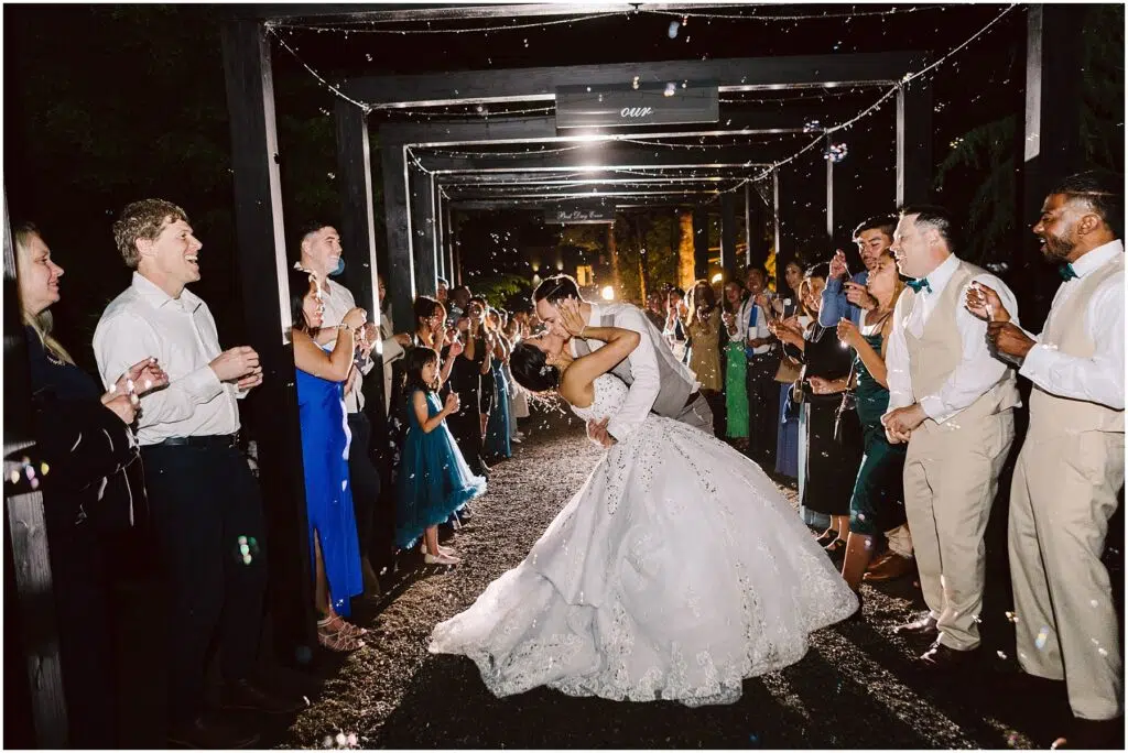 Snohomish Wedding Photographer GSquared Weddings A bride and groom share a kiss under a decorated archway at night, surrounded by applauding guests. The bride wears a white gown, and the groom is in a suit. Bubbles fill the air, adding a magical atmosphere to the scene. Serving Seattle, Snohomish and Orlando