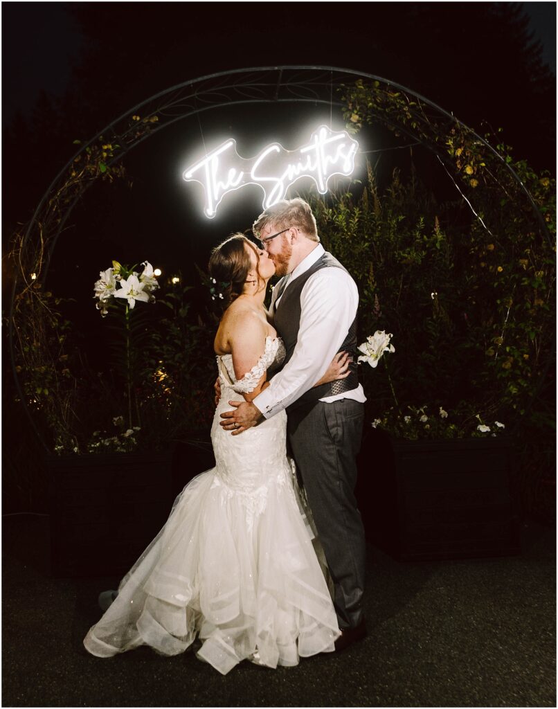 Snohomish Wedding Photographer GSquared Weddings A bride and groom share a kiss under a neon sign reading The Smiths. The bride wears a white gown with lace details, and the groom is in a vest and tie. They are framed by an arch decorated with white flowers and greenery. Serving Seattle, Snohomish and Orlando