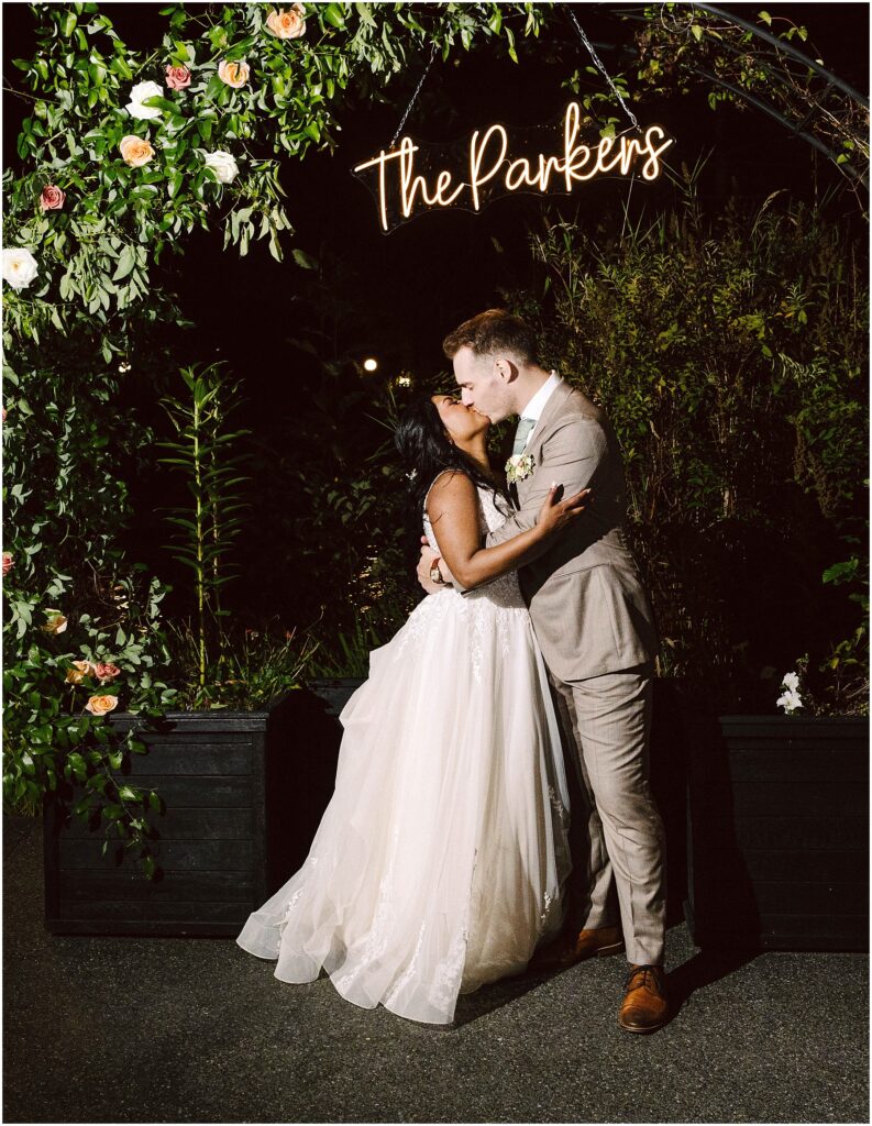 Snohomish Wedding Photographer GSquared Weddings A bride and groom share a kiss under an arch adorned with flowers and greenery. Above them is a neon sign displaying The Parkers. The bride wears a flowing white gown, and the groom is in a light gray suit. It is nighttime. Serving Seattle, Snohomish and Orlando
