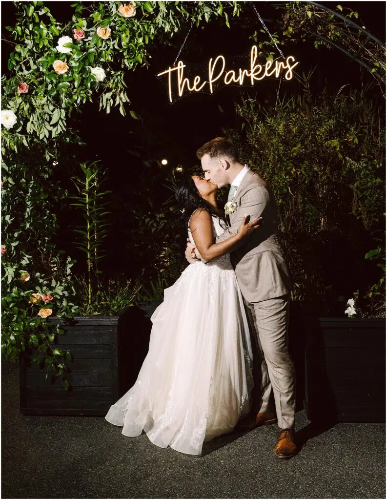 Snohomish Wedding Photographer GSquared Weddings A bride and groom share a kiss under an arch adorned with flowers and greenery. Above them is a neon sign displaying The Parkers. The bride wears a flowing white gown, and the groom is in a light gray suit. It is nighttime. Serving Seattle, Snohomish and Orlando