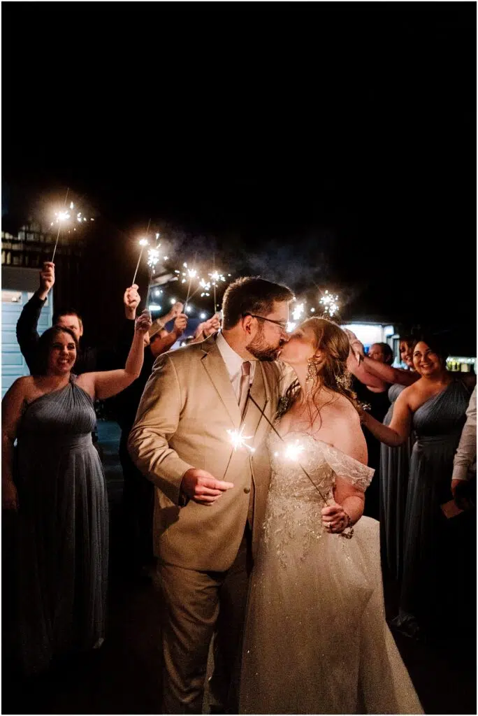 Snohomish Wedding Photographer GSquared Weddings A bride and groom kiss while holding sparklers, surrounded by cheering guests also holding sparklers. The bride wears an off-the-shoulder gown, and the groom is in a light suit. The scene is nighttime, creating a festive, illuminated atmosphere. Serving Seattle, Snohomish and Orlando