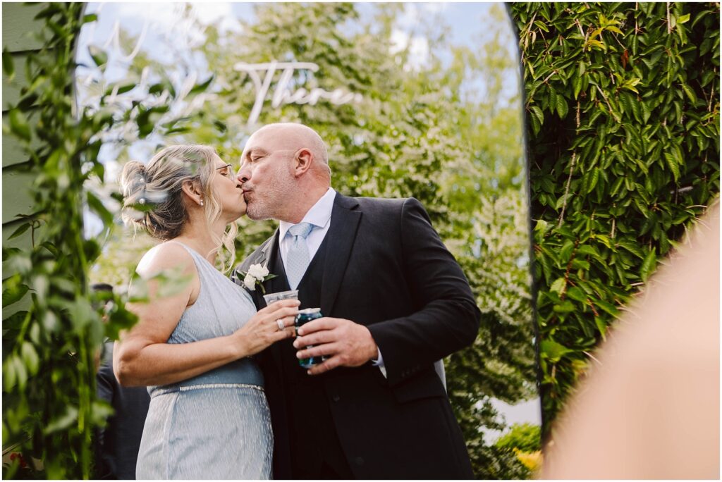 Snohomish Wedding Photographer GSquared Weddings A couple dressed formally shares a kiss in an outdoor setting with lush greenery. The woman holds a drink and wears a light blue dress, while the man, in a suit, holds a beverage can. The background features trees and foliage. Serving Seattle, Snohomish and Orlando