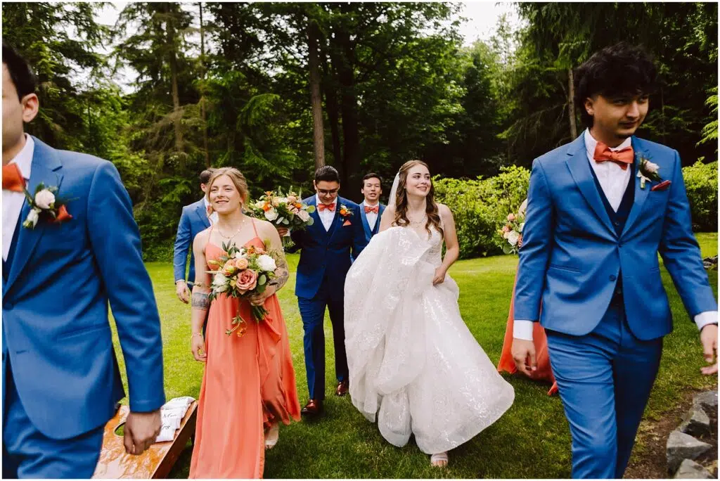 Snohomish Wedding Photographer GSquared Weddings A bride in a white dress walks joyfully with a groom and bridal party through a lush garden. The groomsmen wear blue suits with orange bow ties, and the bridesmaids wear orange dresses, holding bouquets of flowers. Serving Seattle, Snohomish and Orlando