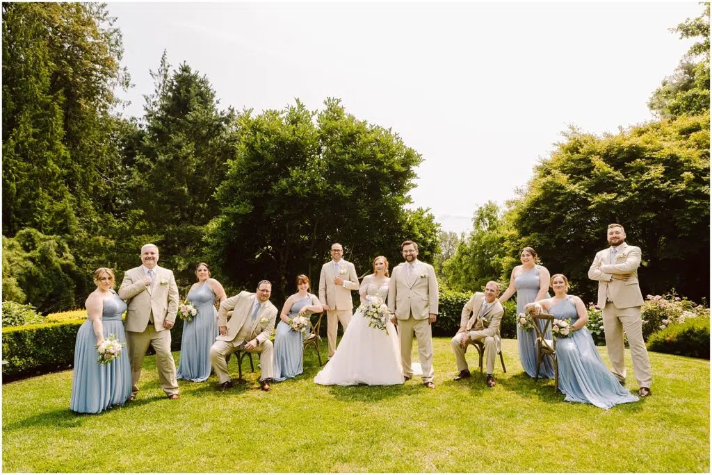 Snohomish Wedding Photographer GSquared Weddings A wedding party poses outdoors on a grassy lawn. The bride and groom stand in the center, surrounded by bridesmaids in light blue dresses and groomsmen in beige suits. Trees and greenery form the backdrop under a sunny sky. Serving Seattle, Snohomish and Orlando