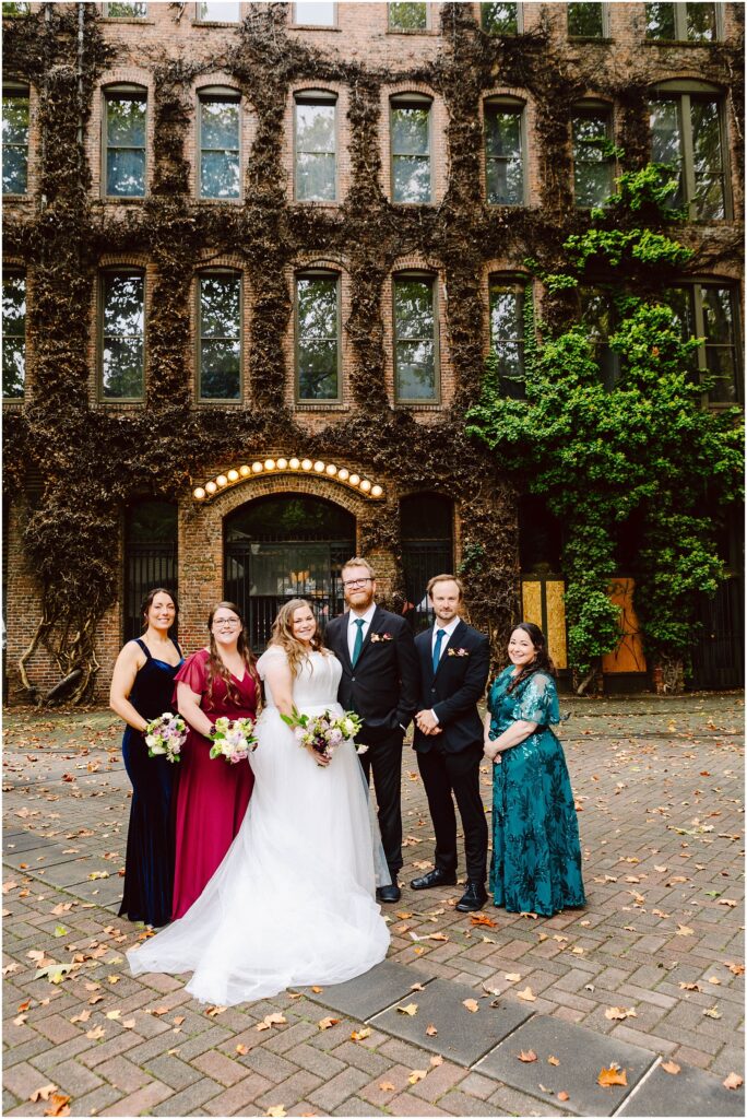 Snohomish Wedding Photographer GSquared Weddings A wedding group poses outdoors in front of a historic brick building with ivy. The bride stands with a groom and four bridesmaids holding bouquets. The scene is set on a cobblestone path with scattered autumn leaves. Serving Seattle, Snohomish and Orlando